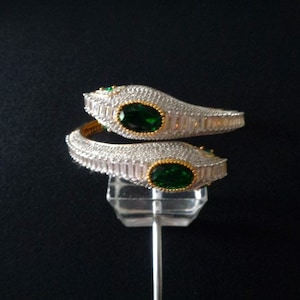 May include: Two silver bracelets in a snake design. Each bracelet has a large oval emerald-green stone in a gold-toned bezel. The bracelets are encrusted with small, clear crystals. The bracelets are displayed on a clear stand against a black background.