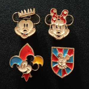 May include: Four enamel pins featuring Mickey Mouse and Minnie Mouse. The pins are gold-toned and feature different designs. One pin shows Mickey Mouse wearing a crown, another shows Minnie Mouse with a red bow, another shows Mickey Mouse in a fleur-de-lis design, and the last pin shows Mickey Mouse in a shield design.