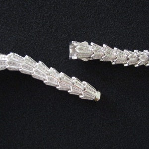 Op de afbeelding: Een zilveren ketting met een delicaat, ingewikkeld ontwerp. De ketting is gemaakt van kleine, met elkaar verbonden stukjes die lijken op een ketting. De ketting is gebroken in twee stukken, waardoor de sluiting zichtbaar is.