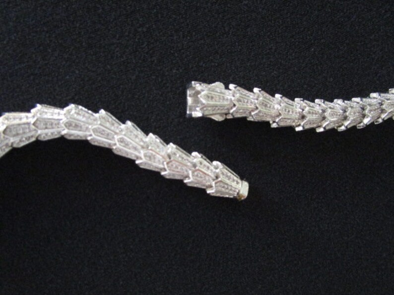 Op de afbeelding: Een zilveren ketting met een delicaat, ingewikkeld ontwerp. De ketting is gemaakt van kleine, met elkaar verbonden stukjes die lijken op een ketting. De ketting is gebroken in twee stukken, waardoor de sluiting zichtbaar is.