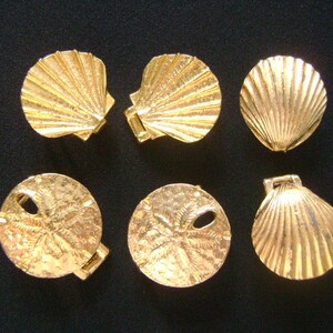 May include: Six gold-colored metal charms in the shape of seashells and sand dollars. The charms are all about 2 inches in diameter.
