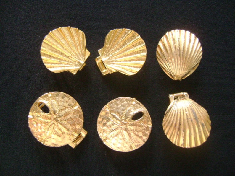 May include: Six gold-colored metal charms in the shape of seashells and sand dollars. The charms are all about 2 inches in diameter.