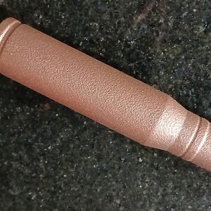 May include: A copper-colored bullet with a textured surface. The bullet has a cylindrical body with a rounded base and a pointed tip. The bullet is lying on a dark, speckled surface.