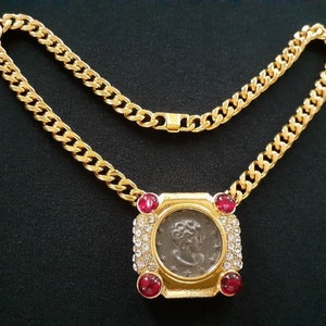 May include: A gold chain necklace with a pendant featuring a silver coin surrounded by clear rhinestones and red gemstones.