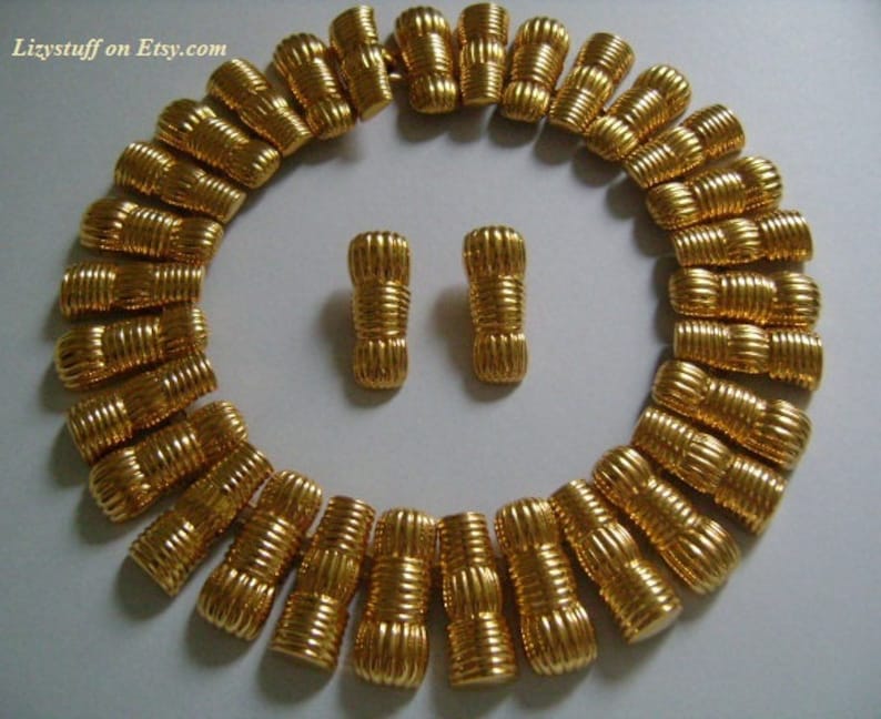 May include: A gold-tone necklace and matching earrings. The necklace is made of a series of linked, textured, cylindrical beads. The earrings are a pair of matching, textured, cylindrical beads.