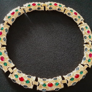 May include: A gold necklace with a repeating pattern of green, red, and blue gemstones. The necklace is made of linked metal pieces with a decorative design.