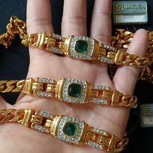 May include: Gold-toned chain bracelets with emerald-colored center stones and surrounding clear crystals. The bracelets have a rectangular design with a buckle-like clasp. The image shows three bracelets, with a dark background.