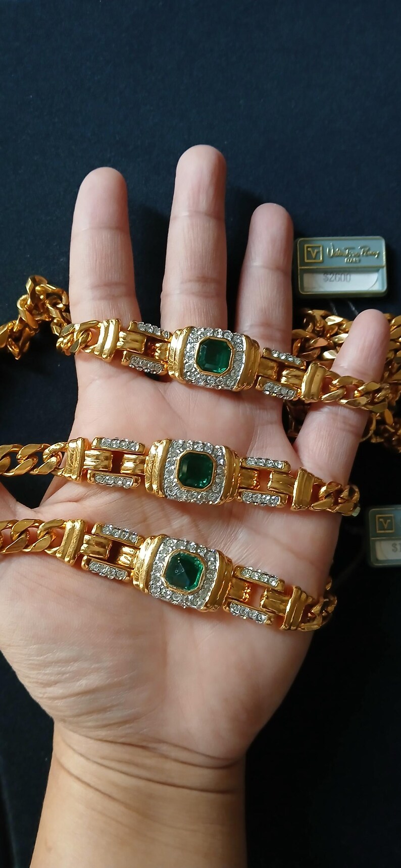 May include: Gold-toned chain bracelets with emerald-colored center stones and surrounding clear crystals. The bracelets have a rectangular design with a buckle-like clasp. The image shows three bracelets, with a dark background.