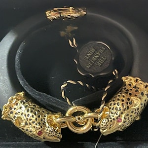 May include: A black leather bracelet with gold-tone leopard head charms. The bracelet is adorned with a black and gold tag that reads "THE FRANKLIN MINT".