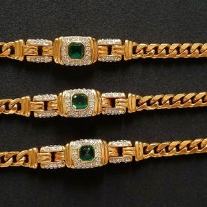 May include: Gold-toned jewelry set featuring a necklace and two bracelets. Each piece has a curb chain design with a central square emerald-colored stone surrounded by smaller clear stones. The jewelry is displayed on a black surface.