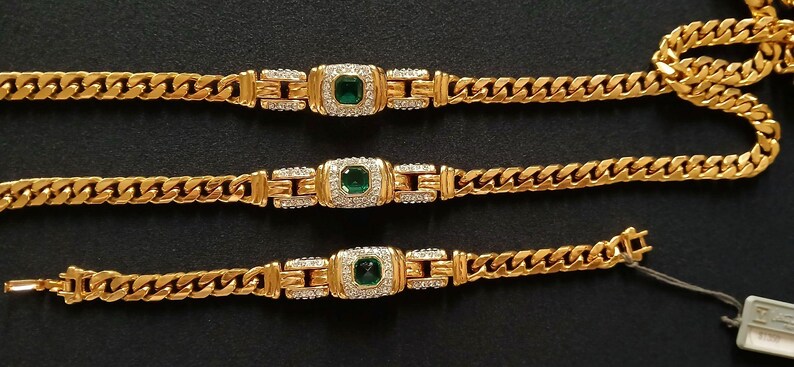 May include: Gold-toned jewelry set featuring a necklace and two bracelets. Each piece has a curb chain design with a central square emerald-colored stone surrounded by smaller clear stones. The jewelry is displayed on a black surface.