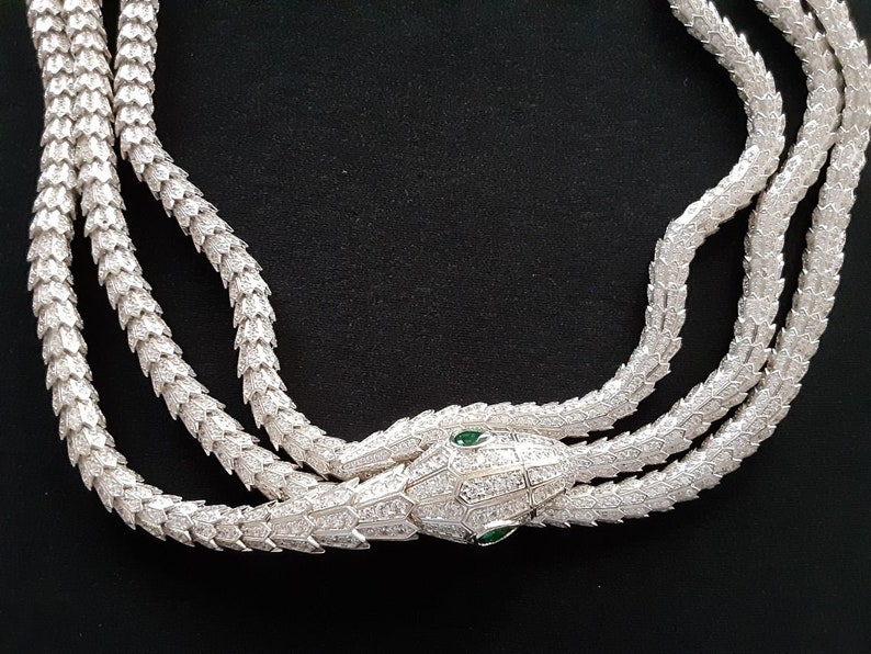 Peut inclure: Un collier de serpent en argent avec un pendentif t&ecirc;te de serpent. La t&ecirc;te de serpent est incrust&eacute;e de pierres claires et a deux yeux verts.
