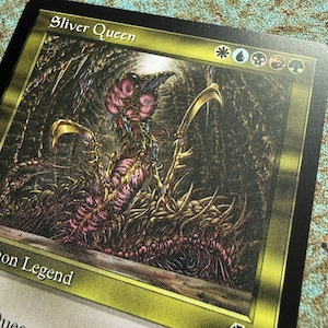 May include: A collectible trading card titled Sliver Queen featuring a dark, detailed illustration of a multi-limbed creature with pink and brown textures. The card has a gold border, five colored mana symbols at the top, and the text Summon Legend.