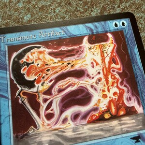 May include: A blue trading card titled Transmute Artifact featuring an illustration of a dark sphere transforming into a glowing, swirling mass of orange, red, and white energy. The card border is blue with two black droplet symbols in the top corner.