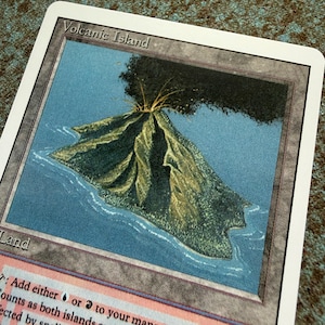May include: A rectangular trading card titled Volcanic Island featuring an illustration of a green volcanic mountain surrounded by blue water. The card has a white border, grey frame, and text at the bottom that reads: Add either blue or red to your mana pool.
