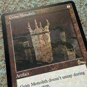 May include: A rectangular trading card titled Grim Monolith featuring a brown border and a central illustration of a tall, weathered stone structure with spires. The card displays the text Artifact and a hammer symbol in the bottom right corner.