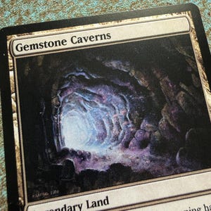 May include: A rectangular trading card titled Gemstone Caverns featuring a dark cavern illustration with glowing purple and blue crystalline rock formations. The card has a white border, black edges, and the text Legendary Land at the bottom.