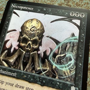 May include: A Magic: The Gathering trading card titled Necropotence featuring a golden skull-faced figure with a spiral marking on its forehead. The artwork displays a swirling light blue energy effect and three black skull mana symbols in the corner.
