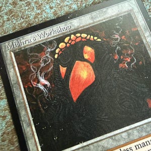 May include: A rectangular trading card with a black border featuring the text Mishra's Workshop at the top. The central illustration depicts a dark, textured figure with glowing orange and yellow highlights, surrounded by wisps of white smoke.