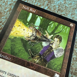 May include: A rectangular trading card titled Null Rod with a brown border. The illustration depicts a person in a purple vest and white sleeves aiming a metallic device that emits a bright yellow energy beam against a green forest background.