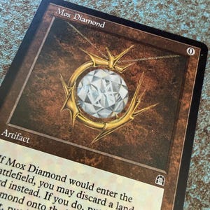 May include: A collectible trading card titled Mox Diamond featuring a large, faceted white diamond set within a gold, thorn-like circular frame. The card has a brown border, the text Artifact, and a zero in the top right corner of the art box.