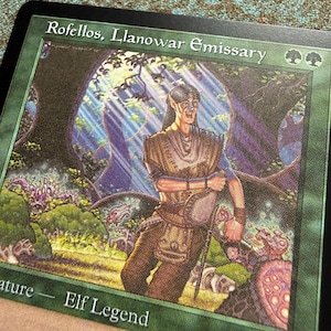 May include: A Magic: The Gathering trading card titled Rofellos, Llanowar Emissary. The card features a green border and an illustration of an elf character in a forest setting with light rays. The text Elf Legend is printed at the bottom.