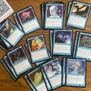 Könnte beinhalten: Eine Sammlung von Magic: The Gathering Sammelkarten, die verschiedene Illustrationen und Texte zeigen. Die Karten haben blaue Ränder mit einzigartigen Kunstwerken und Texten. Ein Deck mit blauen Spielkarten ist ebenfalls zu sehen.