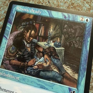 May include: A Magic: The Gathering trading card titled Rhystic Study featuring a blue border and an illustration of a person writing in a book by candlelight. The card displays the text Rhystic Study at the top and Enchantment at the bottom.