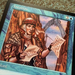 May include: A Magic: The Gathering trading card titled Intuition featuring a character in a brown jacket and head wrap holding a parchment with red symbols. The card has a blue border, a mana cost of 2 and one blue symbol, and a stone archway.