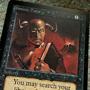 May include: A Magic: The Gathering trading card titled Demonic Tutor, featuring a horned figure holding a dark book against a red volcanic background. The card text includes the name, mana cost, type Sorcery, and partial game instructions.