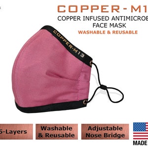 May include: A pink face mask with black trim and a copper-colored logo that reads "COPPER-M13". The mask has an adjustable nose bridge and is made with 5 layers. The text "COPPER INFUSED ANTIMICROBIAL FACE MASK WASHABLE & REUSABLE" is printed above the mask. The text "5-Layers", "Washable & Reusable", "Adjustable Nose Bridge", and "MADE IN USA" are printed below the mask.