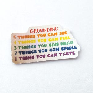 May include: A sticker with the word "GROUNDING" in rainbow colors. Below the word are the phrases "5 THINGS YOU CAN SEE", "4 THINGS YOU CAN FEEL", "3 THINGS YOU CAN HEAR", "2 THINGS YOU CAN SMELL", and "1 THING YOU CAN TASTE".