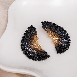 Könnte beinhalten: Ein Paar schwarze und goldene Perlenohrringe in Fächerform. Die Ohrringe bestehen aus kleinen Perlen, die in einem Farbverlauf angeordnet sind, der unten mit Gold beginnt und oben in Schwarz übergeht.