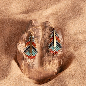 May include: A pair of gold dangle earrings with blue, red, and black beaded fringe. The earrings are displayed on a brown rock.