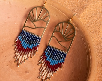 May include: A pair of gold-toned earrings with a sun and mountain design. The earrings are beaded with blue, red, and cream colored beads.