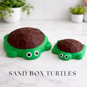 May include: Two green turtle-shaped sandboxes with brown shell lids. The turtles have white eyes and are displayed on a white surface. The text "SAND BOX TURTLES" is at the bottom of the image.