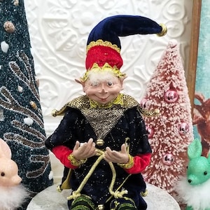 May include: A whimsical elf figurine with a navy blue velvet hat, red trim, and gold accents. The elf is dressed in a black and red outfit with green and black striped leggings. It is sitting on a stone-like pedestal, surrounded by decorative Christmas trees.