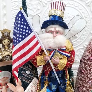 May include: A patriotic Uncle Sam fairy figurine with an American flag. The figure wears a red, white, and blue outfit with gold accents, a top hat, and glittery wings. Other decorative items are in the background.