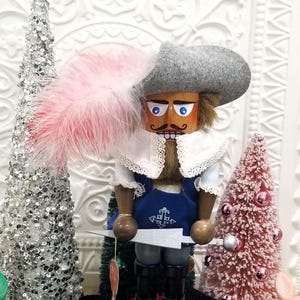 May include: A wooden nutcracker figurine dressed in a blue and white outfit, gray hat, and a pink feather. The nutcracker holds a silver sword and stands on a red base with the word "Aramis".