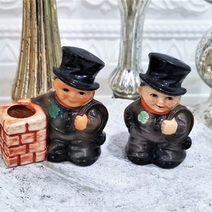 May include: Two small ceramic figurines of chimney sweeps wearing black hats and holding soot bags. The figurines are standing on a white surface next to a small brick chimney.
