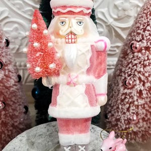 May include: A decorative Christmas nutcracker figurine in shades of pink, white, and silver, holding a pink tree with white pearl accents. The figurine has a detailed face and is wearing a pink and white uniform. A small pink deer ornament is next to the nutcracker.