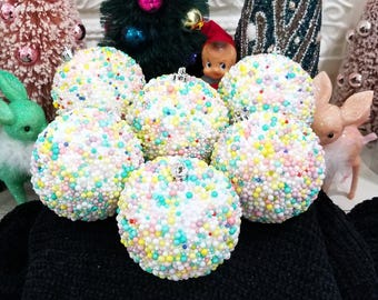 May include: A collection of six round Christmas ornaments covered in small, colorful beads. The ornaments are white with pastel-colored beads in shades of pink, yellow, blue, and green. Silver metal caps are at the top of each ornament.