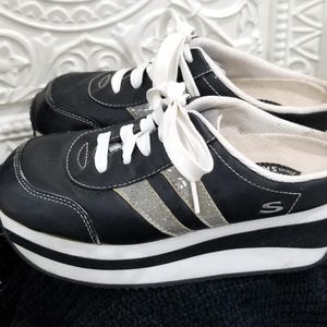 May include: Black platform sneakers with white laces and a silver stripe. The sneakers have a thick white and black sole and the brand name "Skechers" is visible on the side.