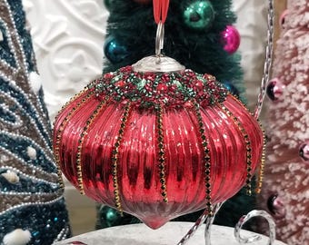 Puede incluir: Una bola de Navidad roja con un diseño acanalado y detalles de cuentas doradas. La parte superior está adornada con joyas brillantes. Se adjunta una cinta roja para colgarla. La bola se exhibe en un soporte plateado.