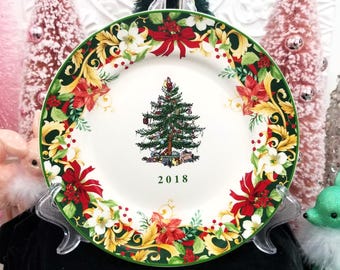 May include: A decorative Christmas plate with a green rim and a white center. The plate features a Christmas tree illustration in the center, surrounded by red bows, poinsettias, and floral designs. The year "2018" is printed below the tree.