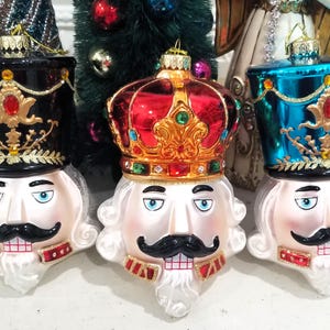 May include: Three decorative Christmas ornaments shaped like nutcracker faces. Each has a different hat: black, red crown, and teal. The faces are white with black mustaches, blue eyes, and red and gold accents. These ornaments are festive and add a touch of holiday cheer.