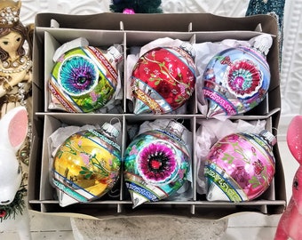 Puede incluir: Un conjunto de seis adornos navideños vintage en una caja dividida. Cada adorno de vidrio presenta una combinación de colores única, que incluye rojo, azul, verde, amarillo y rosa, con diseños florales y geométricos. Los adornos tienen tapas de metal plateado.