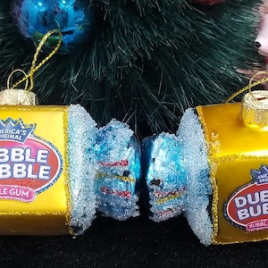 May include: Two Christmas ornaments shaped like DUBBLE BUBBLE bubble gum. The ornaments are yellow with blue and silver glitter accents. Each ornament has the DUBBLE BUBBLE logo. A gold string is attached for hanging.