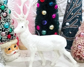 Puede incluir: Una figurita de reno blanca flocada con grandes cuernos. El reno está de pie sobre una superficie blanca frente a una variedad de árboles de Navidad en miniatura.