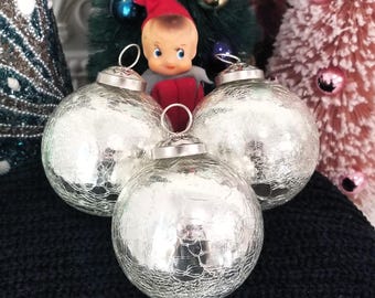 May include: Three silver crackle-glass Christmas ornaments with metal caps and hanging loops. A vintage doll in a red hat and a small Christmas tree are in the background. The ornaments are on a black surface.